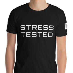 Könnte beinhalten: Ein schwarzes T-Shirt mit kurzen Ärmeln und dem weißen Schriftzug STRESS TESTED in fetter, serifenloser Schrift auf der Brust. Ein kleines quadratisches Logo mit weißem Text ist am linken Ärmel des Kleidungsstücks zu sehen.
