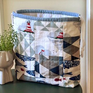 Könnte beinhalten: Ein Stoffbeutel mit Kordelzug und Patchwork-Design, das nautische Motive wie rot-weiße Leuchttürme, blaue Fische und geometrische Muster in Grau-, Blau- und Weißtönen zeigt, mit blauem Stoffrand oben und weißen Kordeln.