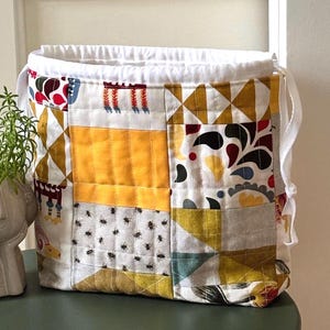 Könnte beinhalten: Ein gesteppter Beutel mit Kordelzug und Patchwork-Design, bestehend aus verschiedenen Mustern wie senfgelben Quadraten, weißem Stoff mit kleinen schwarzen Bienen sowie geometrischen Formen in Rot, Blau und Gelb. Der Beutel hat einen weißen Rand und weiße Bänder.