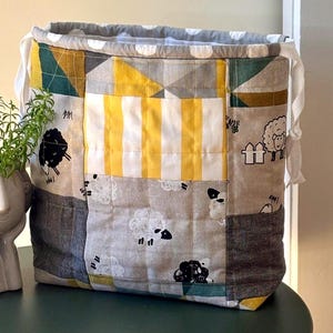 Könnte beinhalten: Ein gesteppter Stoffbeutel mit Kordelzug und einem Patchwork-Design aus Schaf-Illustrationen, gelb-weißen Streifen sowie geometrischen Mustern in Senfgelb, Petrol und Grau. Die Oberkante hat ein grau-weißes Punktefutter mit weißen Bändern.