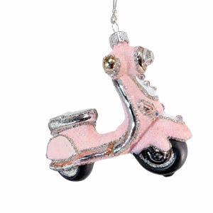 May include: A pink scooter ornament with a glittery finish, silver-toned trim, and black wheels. The decorative piece features a small silver hanging loop at the top, with detailed molded accents on the handlebars, headlight, and seat area.