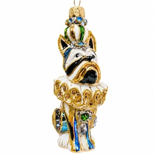 May include: A glass ornament shaped like a dog wearing a gold-trimmed ruff collar and a crown. The dog is white with black markings, blue accents, and gold glitter details. It features a gold hanging loop at the top and decorative jewels.