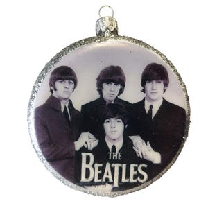 Puede incluir: Una bola de Navidad redonda con una imagen en blanco y negro de The Beatles. Los miembros de la banda visten trajes, con el nombre de la banda en blanco en la parte inferior. La bola tiene un borde plateado con purpurina y una tapa plateada.