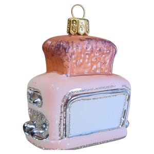 May include: A glass Christmas ornament shaped like a vintage toaster in a soft pink hue. The top features a textured, brownish-orange finish representing bread, while the base and side details are accented with silver glitter and metallic paint.