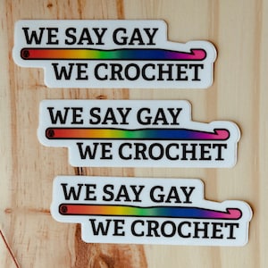 May include: Three white stickers with the text "WE SAY GAY WE CROCHET" in black. Each sticker has a rainbow-colored crochet hook design. The stickers are arranged on a light-colored wooden surface.