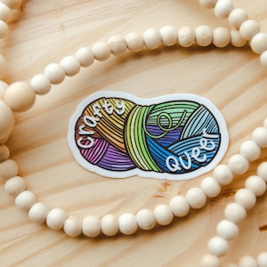 May include: A sticker with the text "crafty queer" over a colorful yarn ball design. The sticker is on a wooden surface, surrounded by a beaded garland.