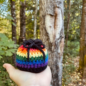 May include: A small rainbow crocheted pouch with a black drawstring. The pouch is being held in a hand in a forest setting.