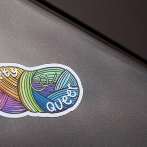 May include: A colorful sticker featuring two yarn balls with the words "crafty queer" in white script. The yarn balls are in shades of orange, yellow, purple, green, and blue. The sticker is on a gray surface.