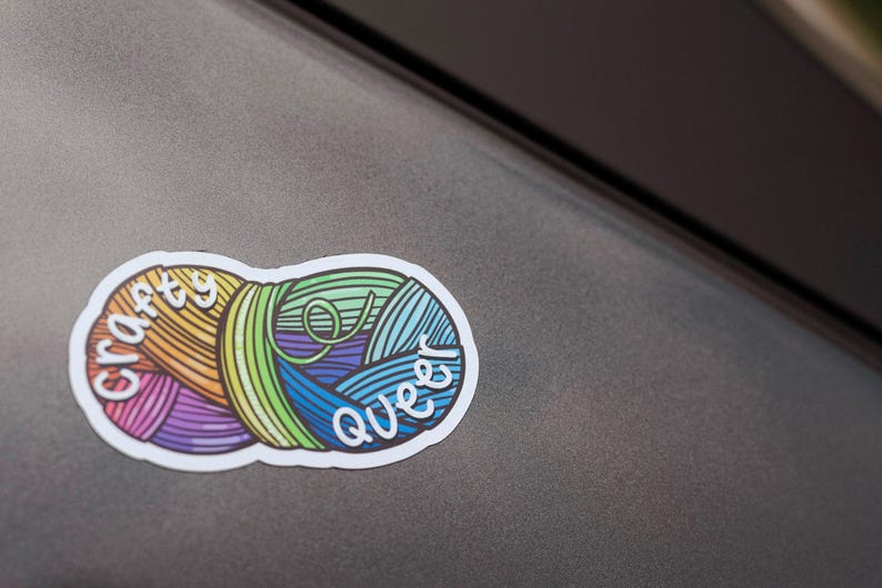 May include: A colorful sticker featuring two yarn balls with the words "crafty queer" in white script. The yarn balls are in shades of orange, yellow, purple, green, and blue. The sticker is on a gray surface.