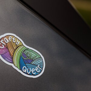 May include: A colorful sticker featuring two yarn balls in rainbow hues. The sticker has the words "crafty queer" in white script. The sticker is on a dark gray surface.
