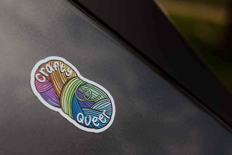 May include: A colorful sticker featuring two yarn balls in rainbow hues. The sticker has the words "crafty queer" in white script. The sticker is on a dark gray surface.
