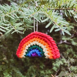 May include: A crocheted rainbow ornament with red, orange, yellow, green, blue, and purple stripes. The rainbow is suspended from a natural twine string against a backdrop of green pine needles. A colorful, handmade decoration.
