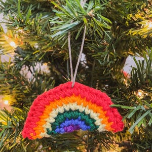 May include: A crocheted rainbow Christmas ornament. The ornament features a red top, followed by bands of orange, yellow, green, blue, and purple. It hangs from a thin, light-colored string against a backdrop of green Christmas tree needles.