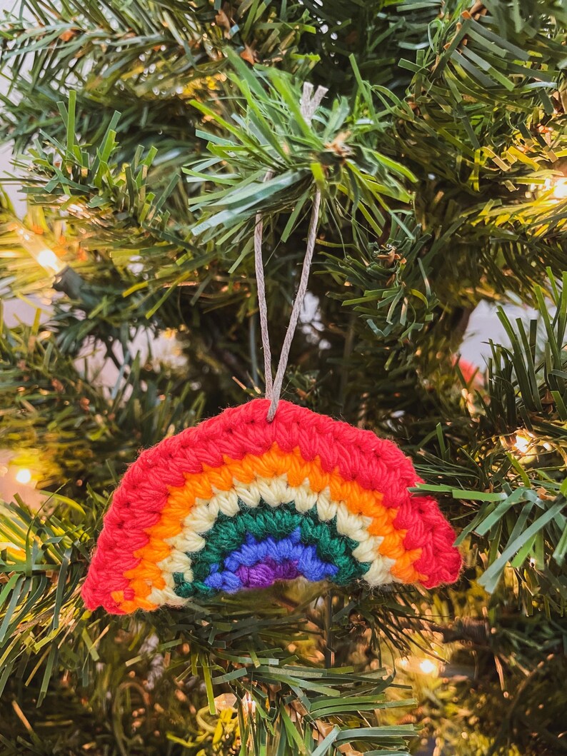 May include: A crocheted rainbow Christmas ornament. The ornament features a red top, followed by bands of orange, yellow, green, blue, and purple. It hangs from a thin, light-colored string against a backdrop of green Christmas tree needles.