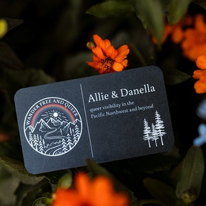 May include: A black business card with rounded corners, featuring the text "Allie & Danella" and "queer visibility in the Pacific Northwest and beyond." The card has a circular logo with mountains, a rainbow, and the words "Wander Free and Queer."