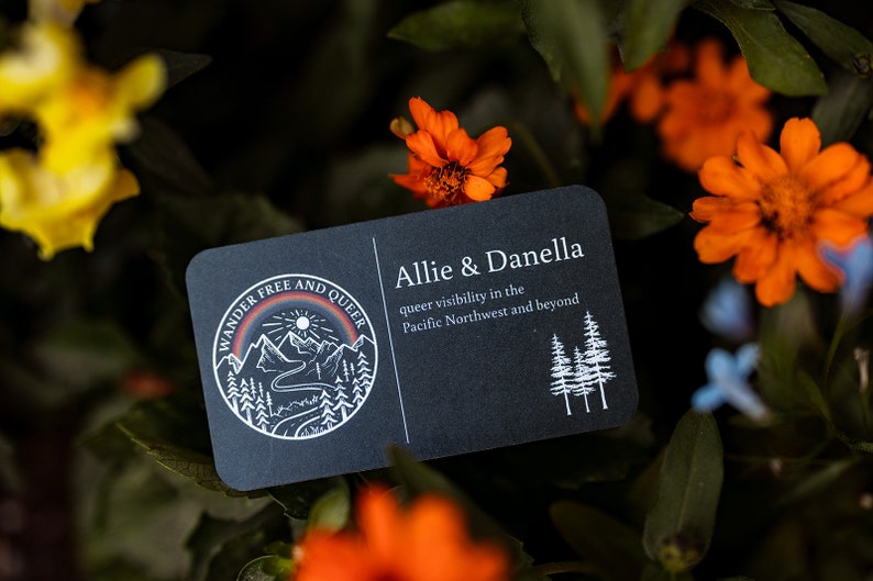 May include: A black business card with rounded corners, featuring the text "Allie & Danella" and "queer visibility in the Pacific Northwest and beyond." The card has a circular logo with mountains, a rainbow, and the words "Wander Free and Queer."