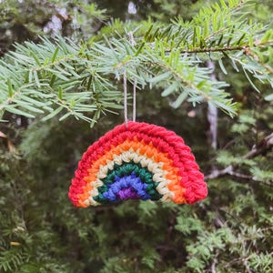 May include: A handmade crochet rainbow ornament with vibrant colors of red, orange, yellow, green, blue, and purple. The ornament is suspended from a natural twine string against a backdrop of green pine needles.