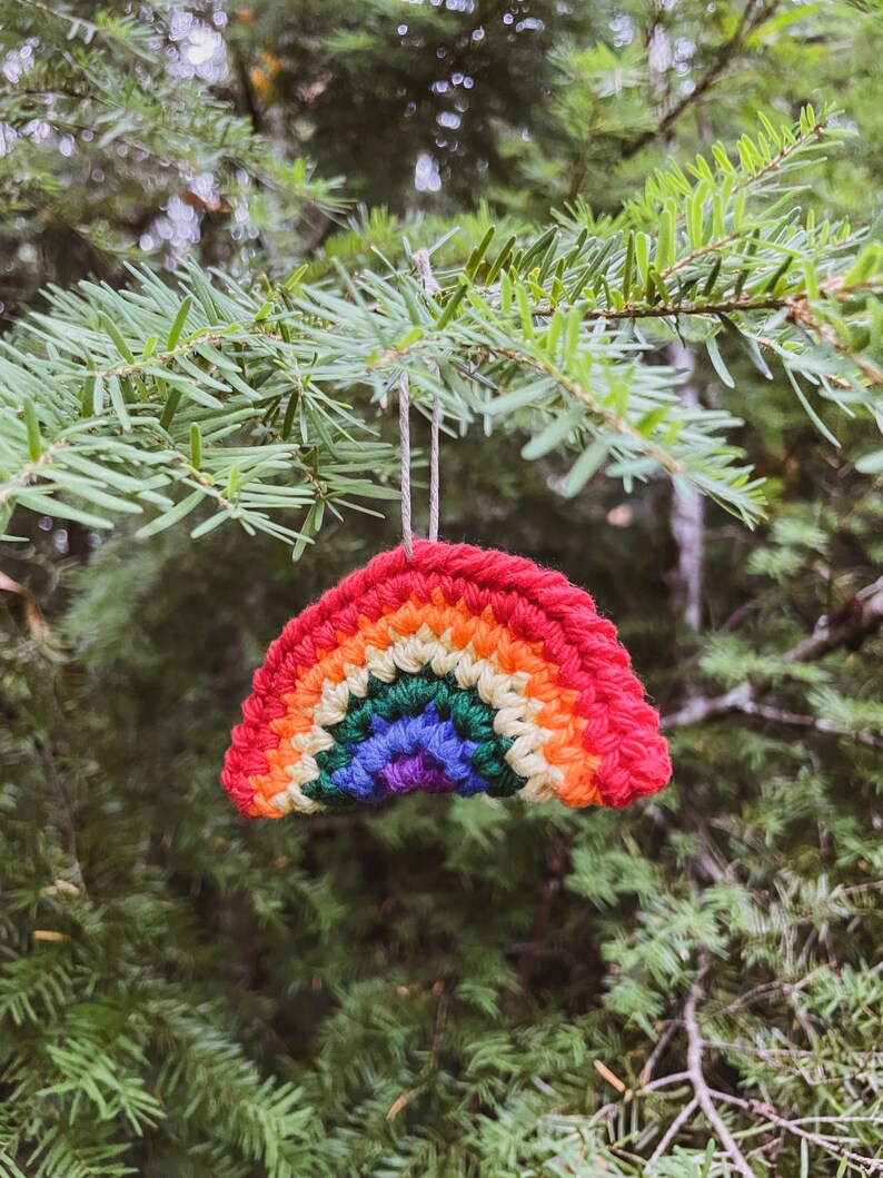 May include: A handmade crochet rainbow ornament with vibrant colors of red, orange, yellow, green, blue, and purple. The ornament is suspended from a natural twine string against a backdrop of green pine needles.