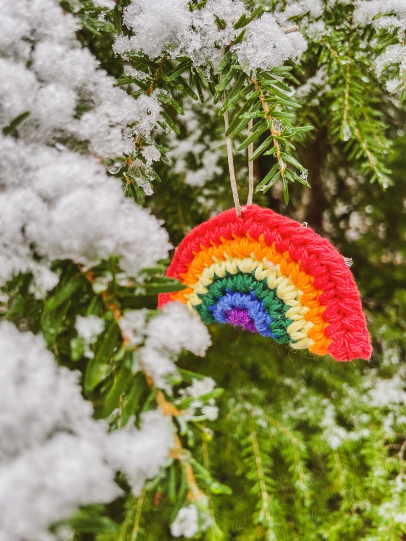 May include: A handmade, crocheted rainbow ornament with red, orange, yellow, green, blue, and purple stripes hangs from a snowy evergreen branch. The colorful decoration is suspended by a natural-colored string, creating a festive winter scene.