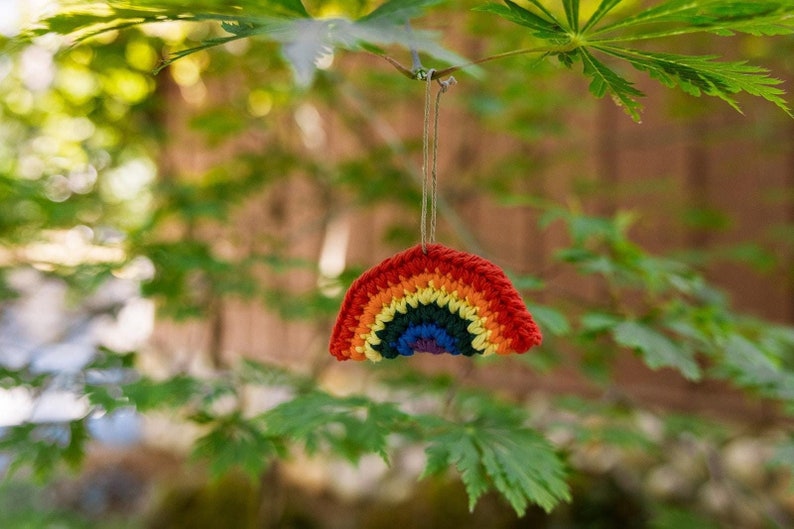 May include: A crocheted rainbow ornament with a red, orange, yellow, green, and blue color scheme. The rainbow is suspended from a natural twine string, hanging against a blurred green background of leaves.