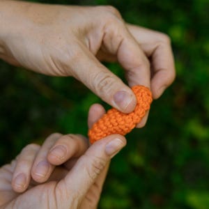 May include: Close-up of an orange crocheted object held between two hands. The object is small, cylindrical, and appears to be handmade. The background is a blurred green, suggesting an outdoor setting.