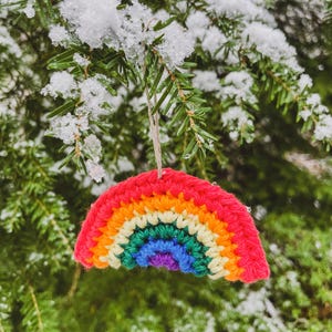 May include: A crocheted rainbow ornament with red, orange, yellow, green, blue, and purple arches hangs from a snowy evergreen branch. The colorful decoration is suspended by a natural twine string, creating a cheerful winter scene.