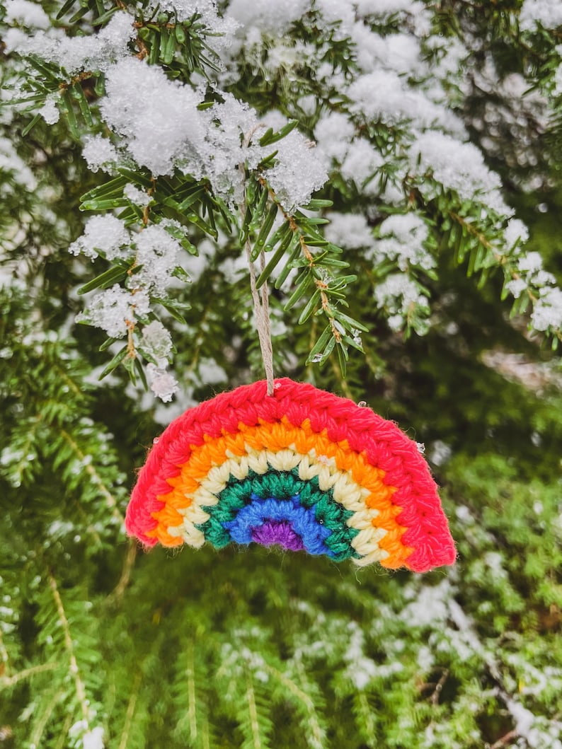 May include: A crocheted rainbow ornament with red, orange, yellow, green, blue, and purple arches hangs from a snowy evergreen branch. The colorful decoration is suspended by a natural twine string, creating a cheerful winter scene.