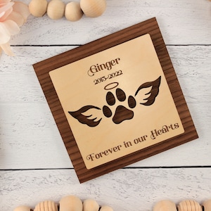 May include: A wooden plaque with a light brown background and a dark brown border. The plaque has a paw print with angel wings and a halo above it. The text "Ginger 2013-2022 Forever in our Hearts" is engraved on the plaque.