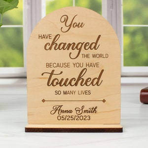 May include: Wooden sign with the text "You have changed the world because you have touched so many lives." The sign also includes the name "Anna Smith" and the date "05/25/2023".
