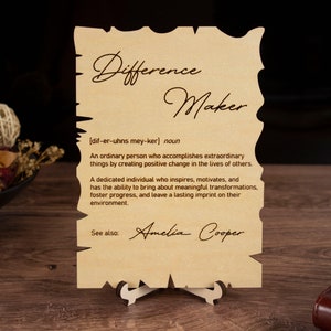 May include: A wooden plaque with a definition of the word "Difference Maker" and the name "Amelia Cooper" written below. The plaque is on a small wooden stand.