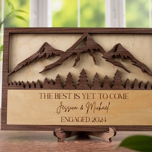 May include: A wooden sign with a mountain and forest design. The text on the sign reads "The best is yet to come Jessica & Michael Engaged 2024".