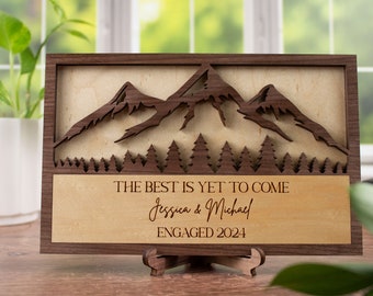 May include: A wooden sign with a mountain and forest design. The text on the sign reads "The best is yet to come Jessica & Michael Engaged 2024".
