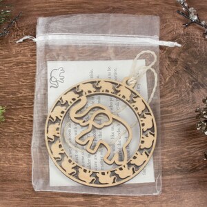 May include: A wooden ornament shaped like a circle with a cutout of an elephant in the center. The elephant is surrounded by a ring of smaller elephants. The ornament is packaged in a clear organza bag with a small card that reads "We were known as the predators, He is the elder to scare away the predators, He is one of us, matter who you are, you are part of the circle. Yea, you are my true friend."