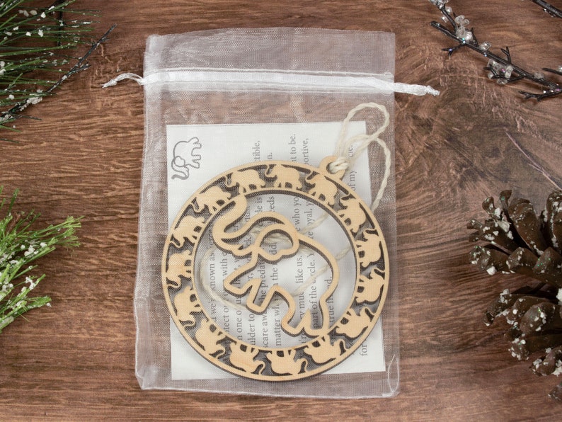 May include: A wooden ornament shaped like a circle with a cutout of an elephant in the center. The elephant is surrounded by a ring of smaller elephants. The ornament is packaged in a clear organza bag with a small card that reads "We were known as the predators, He is the elder to scare away the predators, He is one of us, matter who you are, you are part of the circle. Yea, you are my true friend."