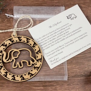 May include: Wooden ornament shaped like a circle with an elephant silhouette inside. The circle is surrounded by smaller elephant silhouettes. The ornament comes with a small card that reads "The Elephant" and includes a poem about elephants.