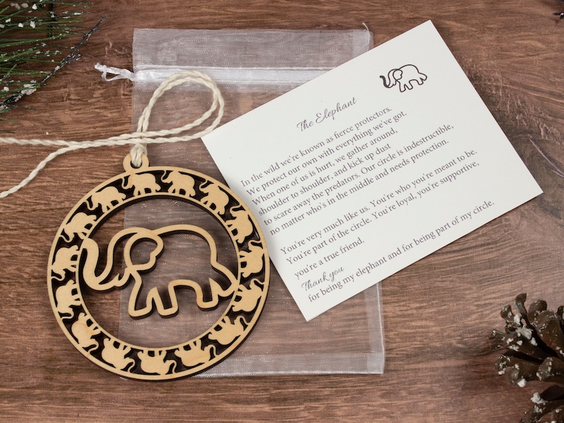 May include: Wooden ornament shaped like a circle with an elephant silhouette inside. The circle is surrounded by smaller elephant silhouettes. The ornament comes with a small card that reads "The Elephant" and includes a poem about elephants.