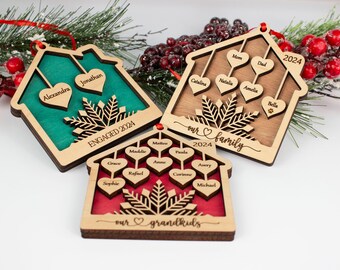 May include: Three wooden house-shaped Christmas ornaments with a snowflake design and personalized names. The ornaments are decorated with red, green, and brown paint. The text on the ornaments reads "Engaged 2024", "Our Family 2024", and "Our Grandkids".