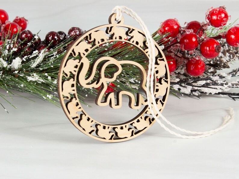 May include: A wooden Christmas ornament shaped like a circle with a large elephant cutout in the center. The ornament is decorated with smaller elephant cutouts around the edge.