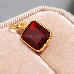 May include: A gold-colored pendant with a rectangular, deep red gemstone. The gemstone is set within a gold frame and has a loop for a chain. The text "JWELZSPARK" is visible on the pendant.