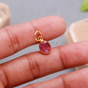 Puede incluir: Un pequeño colgante de forma ovalada con una piedra preciosa facetada de color rosa intenso engastada en un bisel de metal dorado. El colgante incluye una anilla circular en la parte superior para su sujeción, con un acabado pulido y un tono rojo vibrante y translúcido.