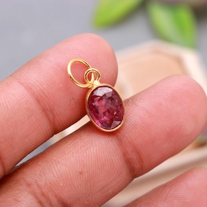 May include: A gold-colored pendant with a vibrant, oval-shaped, red gemstone. The pendant has a small loop at the top for attaching to a chain or bracelet. The gemstone is set in a gold bezel.