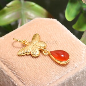 May include: A gold-colored pendant featuring a butterfly design with detailed wings. A teardrop-shaped, orange-red gemstone hangs below the butterfly. The pendant is set against a soft, peach-colored background.