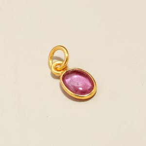 May include: A gold-colored pendant with an oval, pink gemstone. The gemstone is set in a gold bezel and has a polished surface. The pendant has a small loop for attaching to a chain. Jewelry.