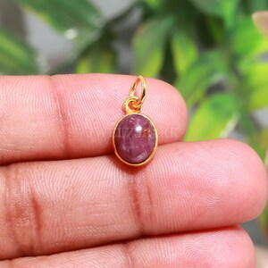 Könnte beinhalten: Ein kleiner ovaler Anhänger mit einem tief rosa-roten Cabochon-Edelstein, der in eine polierte, goldfarbene Metallfassung eingefasst ist. Der Anhänger verfügt über einen kleinen, runden, goldfarbenen Verbindungsring an der Oberseite zur Befestigung.