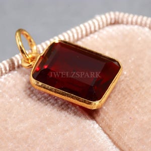 Può includere: Un ciondolo rettangolare con pietra preziosa rosso scuro con castone e anello dorati. Il ciondolo ha il testo "JWELZSPARK" sulla pietra preziosa. Il ciondolo è posto su uno sfondo rosa chiaro e strutturato.