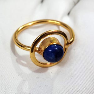 Może przedstawiać: Złoty pierścionek z ciemnoniebieskim, okrągłym kamieniem szlachetnym. Pierścionek ma unikalny design, z kamieniem osadzonym w okrągłej ramie. Pierścionek wykonany jest z błyszczącego, metalicznego materiału.