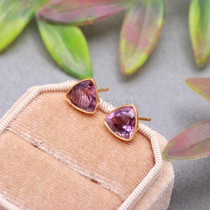May include: A pair of stud earrings featuring triangular-cut purple amethyst gemstones set in gold metal. The earrings are positioned on a soft, light pink velvet display cushion, showcasing the faceted surfaces and the warm metallic tone of the settings.