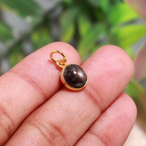 May include: A small oval pendant featuring a dark brown, polished gemstone set in a gold-toned metal bezel. The pendant includes a circular jump ring at the top for attachment, showcasing a smooth, rounded surface and a metallic gold frame.
