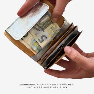 May include: A silver and brown wallet with multiple compartments, holding Euro banknotes and cards. The wallet is open, revealing its accordion-style design. The text on the image reads "ZIEHHARMONIKA-PRINZIP - 4 F&Auml;CHER UND ALLES AUF EINEN BLICK."