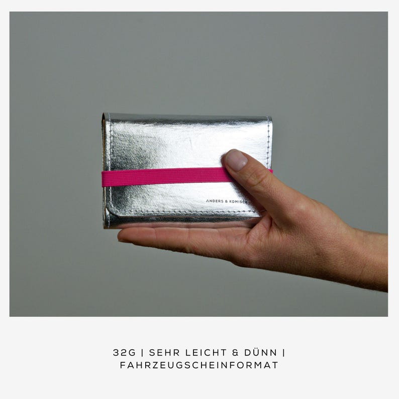 May include: A silver wallet with a pink elastic band, held in a hand. The wallet has a textured metallic finish and the text "ANDERS & KOMISCH" is visible. The bottom of the image has the text "32G | SEHR LEICHT & D&Uuml;NN | FAHRZEUGSCHEINFORMAT".